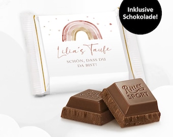 May include: A white chocolate bar wrapper with a pink and gold rainbow design. The wrapper has the text "Lilia's Taufe" and "SCHÖN, DASS DU DA BIST!"  The wrapper is accompanied by two Ritter Sport chocolate bars.