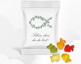 May include: A white pouch with a green leafy fish design and the text "Schön, dass du da bist!"  The pouch is open and there are five colorful gummy fish on a white surface.