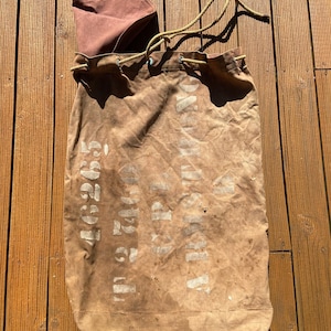 May include: A vintage brown canvas duffel bag with a drawstring closure and a brown interior. The bag has white stencilled text and numbers. The drawstring is a light green colour. The bag is sitting on a wooden surface.