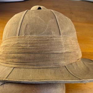 May include: A tan pith helmet featuring a structured dome crown with a small top vent and a wide, downward-sloping brim. The hat is wrapped with a wide, textured fabric band that has visible frayed edges and loose threads across its surface.