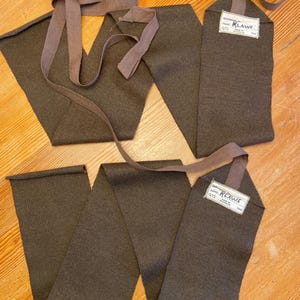 May include: Two olive-drab fabric leg wraps with attached straps and tags. The tags read "KLAWS" and include additional text. The wraps are laid out on a wooden surface.