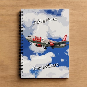 May include: Spiral-bound notebook with a Jet airplane design against a blue sky and clouds. The notebook has the text "Add a Name" and "Your text here". The cover is on a wooden surface.