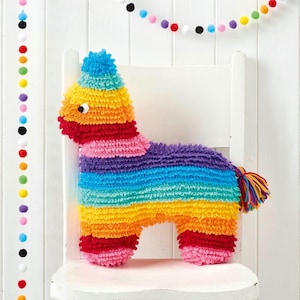 May include: A handmade, colorful, crocheted donkey piñata with a rainbow striped body, a blue and green pom-pom hat, and a multicolored tail. The piñata is sitting on a white chair, with a garland of colorful pom-poms in the background.