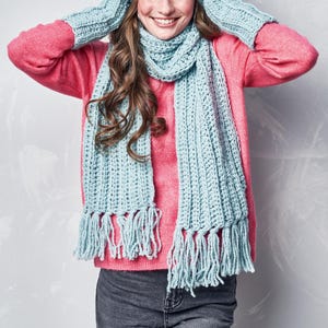 May include: A light blue crochet scarf, hat, and mittens set with fringe. The set is displayed against a pink sweater and gray background. The scarf is long and rectangular, with a ribbed texture. The mittens and hat have a matching ribbed design.