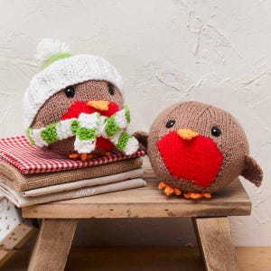 May include: Two knitted robin bird ornaments. One bird wears a white hat with a green pom-pom and a green and white striped scarf. The other bird has a red breast. Both have black eyes and orange feet.
