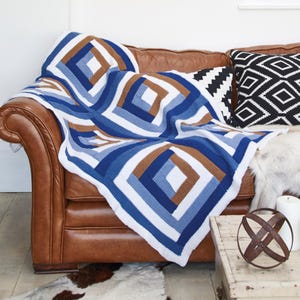 May include: A crochet blanket featuring a geometric pattern of concentric squares in shades of blue, white, and brown. The textile displays a repeating square motif constructed with visible yarn stitches, creating a textured surface across the entire piece.