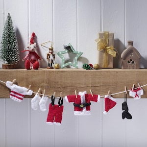 Può includere: Un'esposizione natalizia festosa con un albero in miniatura, ornamenti e una ghirlanda di minuscoli vestiti natalizi lavorati a maglia. L'abbigliamento comprende un costume da Babbo Natale, un cappello e biancheria intima, tutti appesi a un filo.