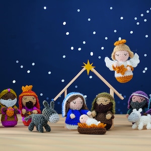 Può includere: Un presepe all'uncinetto con una stella, un angelo e figure di Maria, Giuseppe, Gesù bambino, un asino, una pecora e tre re magi. La scena è ambientata su uno sfondo blu scuro con punti bianchi.