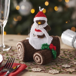 May include: A knitted Christmas decoration featuring a brown log with a knitted robin wearing a red hat and white scarf. The robin is perched on the log, which has a smaller log beside it, and is decorated with holly and berries.