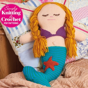 Puede incluir: Una muñeca de sirena de crochet con trenzas largas rubias, un top de bikini morado y una cola a rayas azul y verde. La muñeca está tumbada en una cama con una colcha estampada.