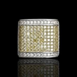 May include: A rectangular ring with a silver band and a surface covered in small, yellow and clear stones. The stones are arranged in a grid pattern, creating a sparkling effect. The ring is set against a black background.