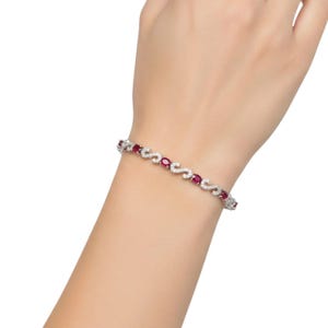 May include: A silver-toned bracelet featuring a repeating pattern of oval-cut red rubies alternating with S-shaped links encrusted with small, clear white stones. The jewellery piece wraps around the wrist, showcasing a metallic finish and vibrant red gems.