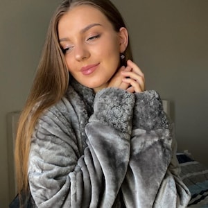 May include: A gray plush robe with a shawl collar and a matching belt. The robe has a soft, textured appearance and is worn by a person. The sleeves have a faux fur trim. The background is a neutral color.