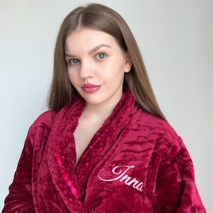 May include: A burgundy plush robe with the name "Inna" embroidered in white script. The robe has a shawl collar and a soft, textured appearance. The image is a close-up of the robe.