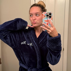 May include: A navy blue plush robe with a shawl collar and a matching belt. The robe has the embroidered initials "M & M" on the chest. The robe is made of a soft, velvety material.