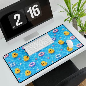 May include: A rectangular desk mat featuring a repeating pattern of yellow rubber ducks, colorful striped life preservers, and small pink and white flowers set against a bright blue, rippling water background with a black stitched border.
