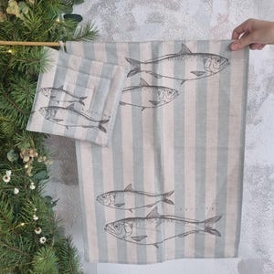 Può includere: Un set di biancheria da cucina con un motivo a righe in azzurro e beige, decorato con illustrazioni di pesci. Il set comprende un canovaccio e un sottobicchiere quadrato. La parola "Herring" è stampata sul canovaccio.