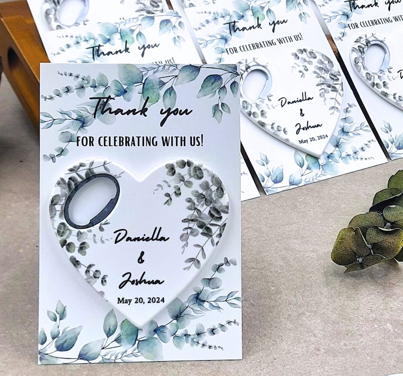 Może przedstawiać: Biały otwieracz do butelek w kształcie serca z wzorem liści eukaliptusa. Otwieracz do butelek ma czarny metalowy pierścień i tekst "Daniella & Joshua May 20, 2024".