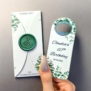 May include: A white card with a green wax seal and the words "Thank You". A bottle opener with the text "Claudia's 60th Birthday" and botanical accents is held in a hand. The date 09/10/2025 is printed on the bottle opener.