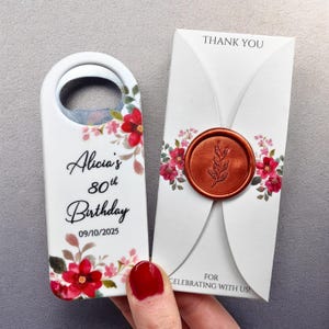 May include: A white bottle opener with a floral design and the text "Alicia's 80th Birthday 09/10/2025". Next to it is a white card with a floral design, a copper seal, and the text "Thank You" and "For Celebrating With Us!"