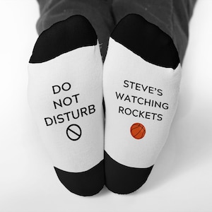 May include: White socks with black heels and toes. One sock reads "DO NOT DISTURB" with a no-entry symbol. The other sock says "STEVE'S WATCHING ROCKETS" with an orange basketball graphic. The socks are on a person's feet.
