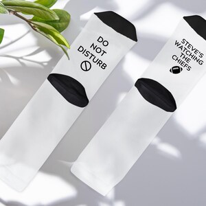 May include: Two white socks with black accents. One sock reads "DO NOT DISTURB" with a no-entry symbol. The other sock says "STEVE'S WATCHING THE CHIEFS" with a football graphic. The socks are on a white surface.