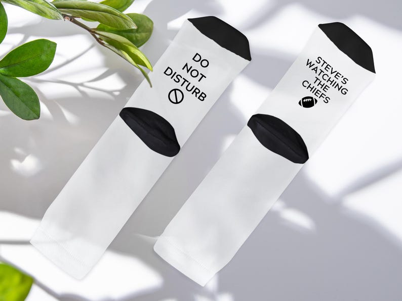 May include: Two white socks with black accents. One sock reads "DO NOT DISTURB" with a no-entry symbol. The other sock says "STEVE'S WATCHING THE CHIEFS" with a football graphic. The socks are on a white surface.