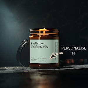 May include: A lit candle in an amber glass jar with a light green label. The label reads: Smells like Wellfleet, MA, Infused with Cape Cod breezes, famous oysters, & the timeless charm of Wellfleet. A hand-drawn arrow points to the label with the text PERSONALISE IT.