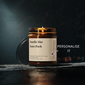 May include: A brown glass jar candle with a lit wick and a cream-colored label. The label features the text Smells like Estes Park, Infused with Rocky Mountain views, elk-filled streets, & the charm of Estes Park. A black lid sits beside the jar.