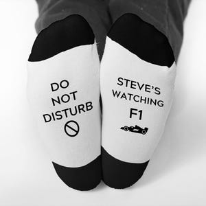 May include: White socks with black heels and toes. One sock reads "DO NOT DISTURB" with a no-entry symbol, the other says "STEVE'S WATCHING F1" with a race car graphic. The socks are worn by a person.