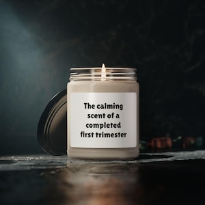 May include: A beige candle with a white label that reads "The calming scent of a completed first trimester".