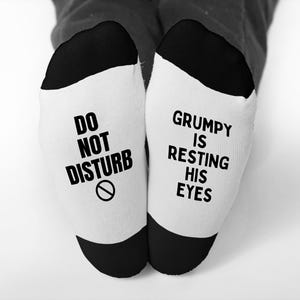 May include: A pair of white socks with black heels and toes. One sock reads "DO NOT DISTURB" with a no-entry symbol, and the other reads "GRUMPY IS RESTING HIS EYES" in black block letters. The socks are worn by a person.