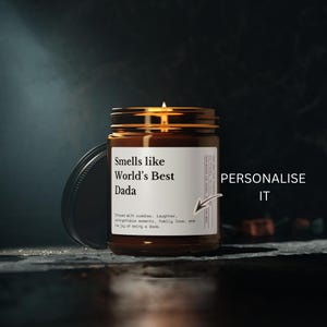 Könnte beinhalten: Ein braunes Kerzenglas aus Glas mit einem weißen Etikett, das den schwarzen Text Smells like World's Best Dada sowie kleineren Text über Kuscheln und Lachen enthält. Ein kleiner Pfeil zeigt auf das Etikett mit dem Text PERSONALIZE IT.