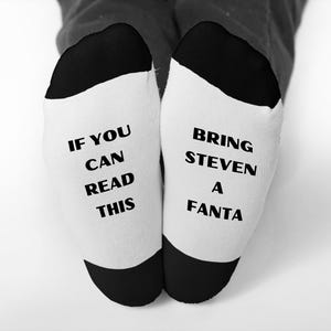 May include: White socks with black heels and toes. One sock reads "IF YOU CAN READ THIS" in black text. The other sock reads "BRING STEVEN A FANTA" in black text. The socks are on a person's feet.