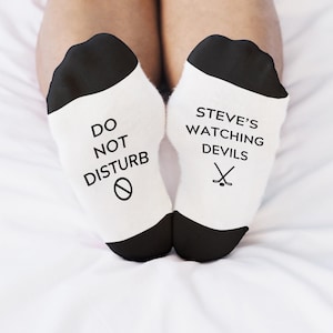 May include: A pair of white socks with black heels and toes. One sock reads "DO NOT DISTURB" with a no-entry symbol. The other sock says "STEVE'S WATCHING DEVILS" with a hockey stick and puck graphic.