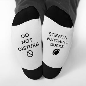 May include: White socks with black heels and toes. One sock reads "DO NOT DISTURB" with a circle and slash symbol. The other sock says "STEVE'S WATCHING DUCKS" with a football graphic. The socks are a novelty item.