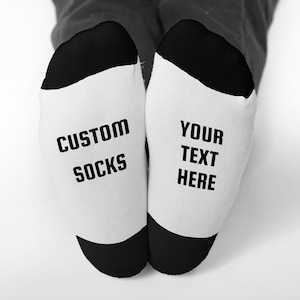 May include: A pair of white socks with black heels and toes. The left sock reads "CUSTOM SOCKS" in black block letters, and the right sock says "YOUR TEXT HERE". The socks are on a white background.