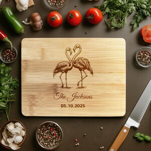 May include: Personalized bamboo cutting board with a laser engraved design of two flamingos facing each other. The text "The Jacksons 05.10.2025" is engraved below the flamingos.