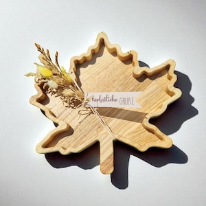 Könnte beinhalten: Ein Ahornblatt-Tablett aus Holz mit hellbraunem Finish. Das Tablett hat ein kleines Bündel getrockneter Blumen und ein Etikett mit der Aufschrift "herbstliche GRÜSSE". Das Tablett befindet sich auf einer weißen Oberfläche.