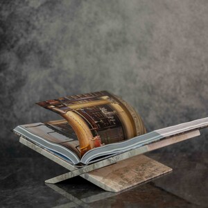 K&ouml;nnte beinhalten: Ein B&uuml;cherst&auml;nder aus Holz mit brauner und beigefarbener Oberfl&auml;che. Ein Buch mit einem bunten Bild eines Raumes liegt auf dem St&auml;nder.