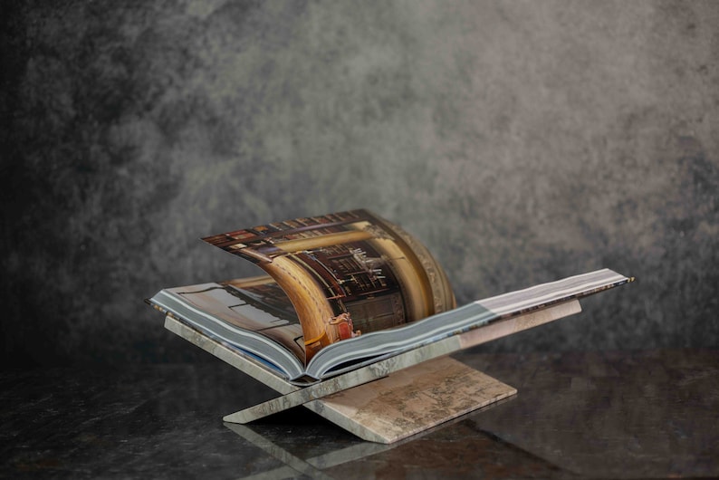 K&ouml;nnte beinhalten: Ein B&uuml;cherst&auml;nder aus Holz mit brauner und beigefarbener Oberfl&auml;che. Ein Buch mit einem bunten Bild eines Raumes liegt auf dem St&auml;nder.