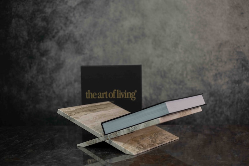 K&ouml;nnte beinhalten: Ein Buch mit dem Titel "the art of living" wird auf einem dekorativen St&auml;nder aus Marmor pr&auml;sentiert. Das Buch ist geschlossen und hat einen hellblauen Einband mit einem wei&szlig;en R&uuml;cken.
