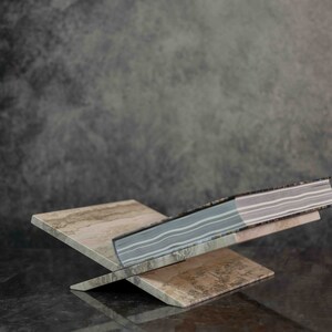 K&ouml;nnte beinhalten: Ein Buch mit einem gestreiften Einband wird auf einem dekorativen Holzbuchst&uuml;tzen pr&auml;sentiert. Der Buchst&uuml;tzen ist aus hellem braunem Holz mit einer strukturierten Oberfl&auml;che. Der Buchst&uuml;tzen ist X-f&ouml;rmig und hat eine rechteckige Basis.