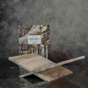 K&ouml;nnte beinhalten: Ein Buch mit dem Titel "Labyrinth" mit einem farbenfrohen Bild eines Geb&auml;udes auf dem Cover wird auf einem hellbraunen Holzbuchst&auml;nder pr&auml;sentiert. Der St&auml;nder besteht aus zwei Holzst&uuml;cken, die im Winkel gekreuzt sind.