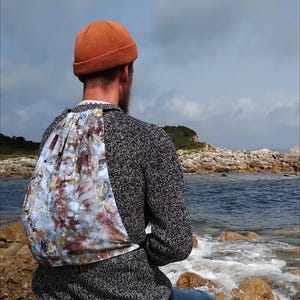 Puede incluir: Una persona con una boina marrón y un suéter gris se sienta en una costa rocosa mirando al mar. Lleva una bolsa de cordón de color tie-dye.