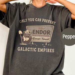 May include: Dark grey t-shirt with the text "ONLY YOU CAN PREVENT" above a sign that reads "ENDOR" and "LUNAR FOREST". Below the sign is the text "GALACTIC EMPIRES". The shirt features a cartoon character with a shovel.