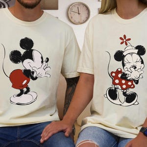 Op de afbeelding: Twee cr&egrave;mekleurige T-shirts met illustraties van Mickey en Minnie Mouse. Mickey draagt een rode korte broek en Minnie een rode jurk met stippen. Beide personages staan in een speelse pose. De shirts worden door twee mensen gedragen, met een blauwe jeans.