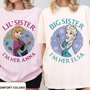 May include: Two t-shirts, one pink and one cream, featuring cartoon characters. The pink shirt says "Lil' Sister I'm Her Anna" with an Anna graphic. The cream shirt says "Big Sister I'm Her Elsa" with an Elsa graphic. The text "Comfort Colors" is at the bottom.