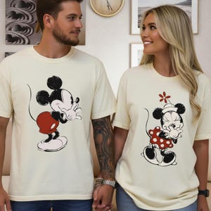 Op de afbeelding: Cr&egrave;mekleurige T-shirts met illustraties van Mickey en Minnie Mouse. Mickey draagt een rode korte broek, terwijl Minnie een rode jurk met stippen draagt. De shirts zijn korte mouwen en worden door twee personen gedragen.