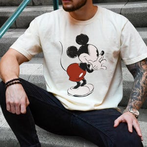 Op de afbeelding: Cr&egrave;mekleurig T-shirt met een Mickey Mouse-afbeelding. Het stripfiguur is afgebeeld in zwart, rood en wit, in een speelse pose. Het shirt is casual kleding, geschikt voor dagelijks gebruik.